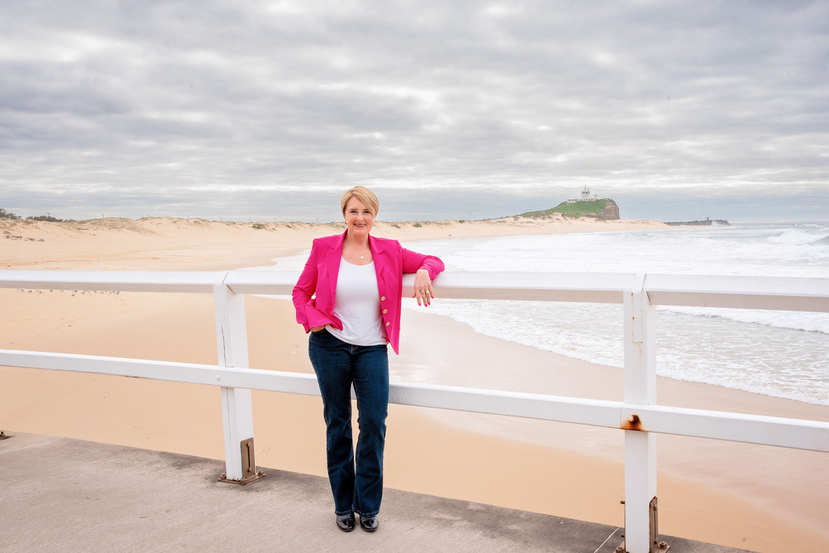 Zoë Routh stands on a beach boardwalk leaning on a white railing, wearing a bright pink blazer over a white top and dark blue jeans, with a sandy beach, ocean waves, and cloudy sky in the background.