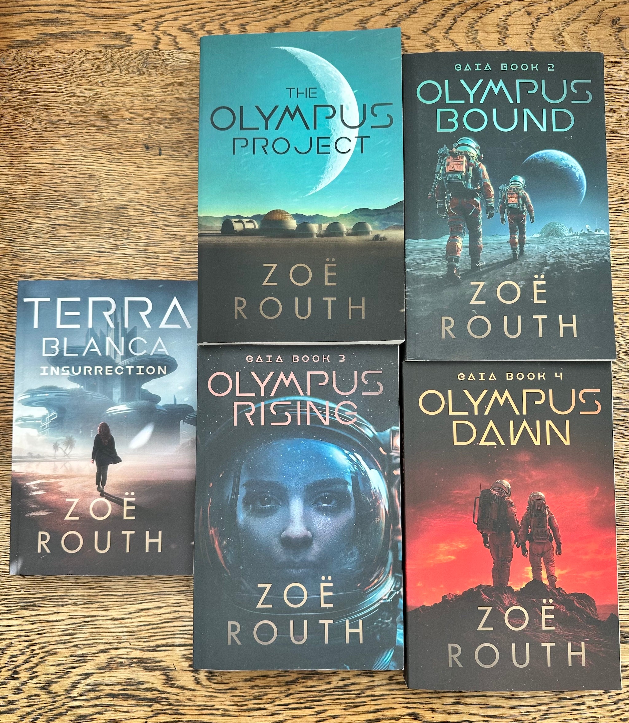 Six paperback books by Zoë Routh arranged on a wooden surface: Terra Blanca Insurrection, The Olympus Project, Gaza Book 2 Olympus Bound, Gaza Book 3 Olympus Rising, Gaza Book 4 Gaza Dawn, and Olympus Dawn. The covers feature space-themed artwork with astronauts, planets, and dramatic sky scenes.