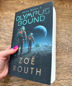A hand with pink nail polish holds a paperback book titled 'Olympus Bound' by Zoë Routh. The book cover features a dark sci-fi illustration of two astronauts walking on a moon-like surface under a large planet, set against a wooden tabletop background.