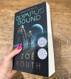 A hand with pink nail polish holds a paperback copy of the book 'Olympus Bound' by Zoë Routh. The book cover is dark with an illustration of two astronauts walking on a moon surface under a planet, with gold and light blue text.