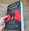 A hand with pink nail polish holds a paperback book titled 'Olympus Dawn' by Zoë Routh. The book cover features a dramatic red and orange sky with two silhouetted astronauts standing on a rocky peak.