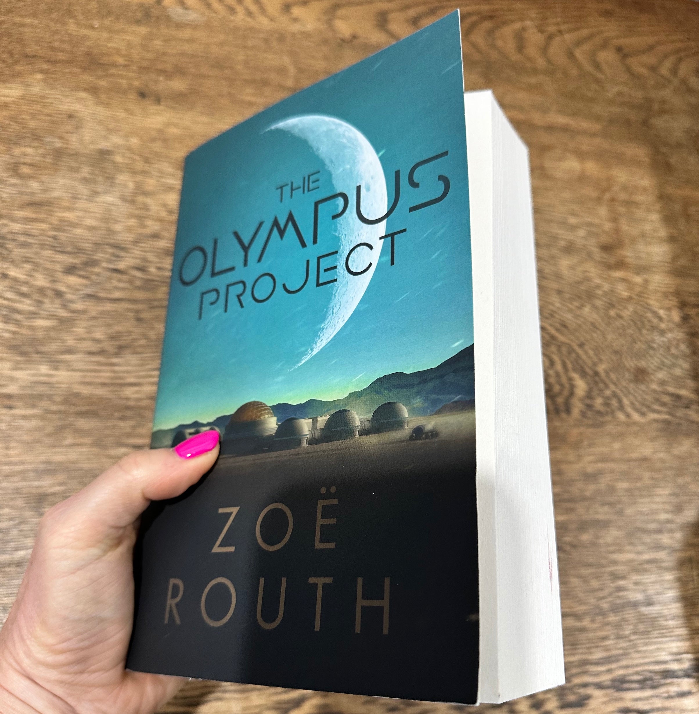 A hand with bright pink nail polish holds a paperback copy of the book 'The Olympus Project' by Zoë Routh, showing the front cover and white pages, set against a wooden tabletop background.
