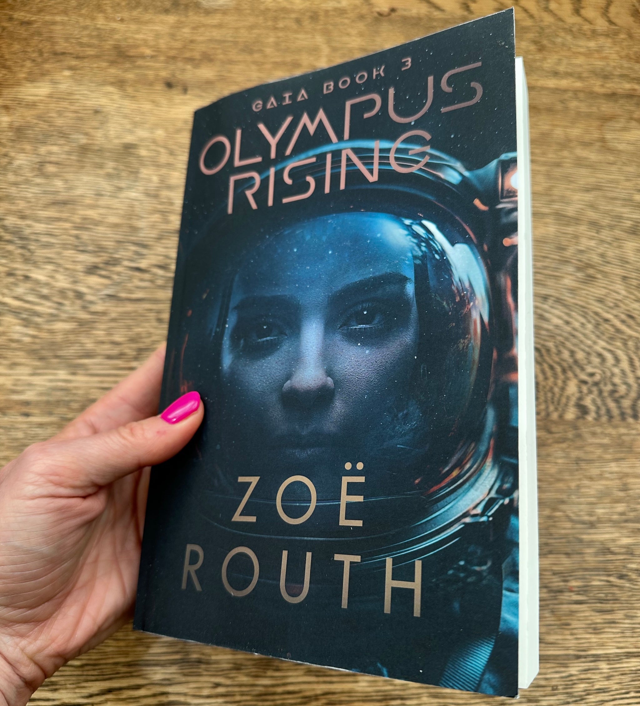 A hand with pink nail polish holds a paperback copy of the book 'Olympus Rising' by Zoë Routh. The book cover features a dark, sci-fi design with an astronaut helmet and the title in a metallic gold font, set against a wooden tabletop background.