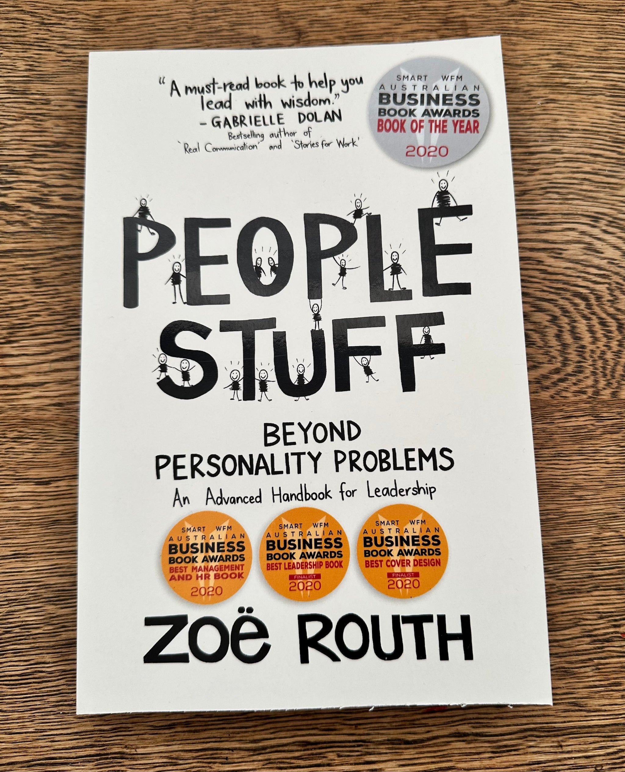 The front cover of the book 'People Stuff: Beyond Personality Problems' by Zoë Routh, displayed flat on a brown wooden surface. The cover is white with bold black typography and small stick-figure illustrations, featuring gold Business Book Awards seals.