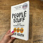 A hand holds the book 'People Stuff: Beyond Personality Problems' by Zoe Roth against a wooden tabletop background. The book cover is white with bold black text and three gold Business Book Awards seals.