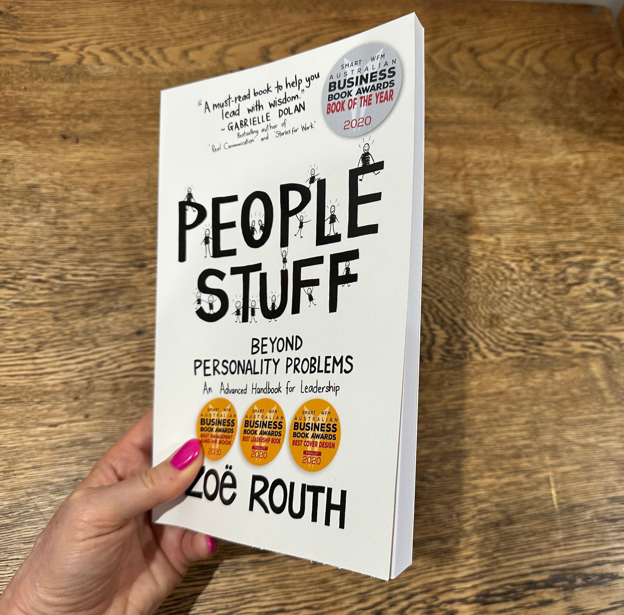 A hand holds the book 'People Stuff: Beyond Personality Problems' by Zoe Roth against a wooden tabletop background. The book cover is white with bold black text and three gold Business Book Awards seals.