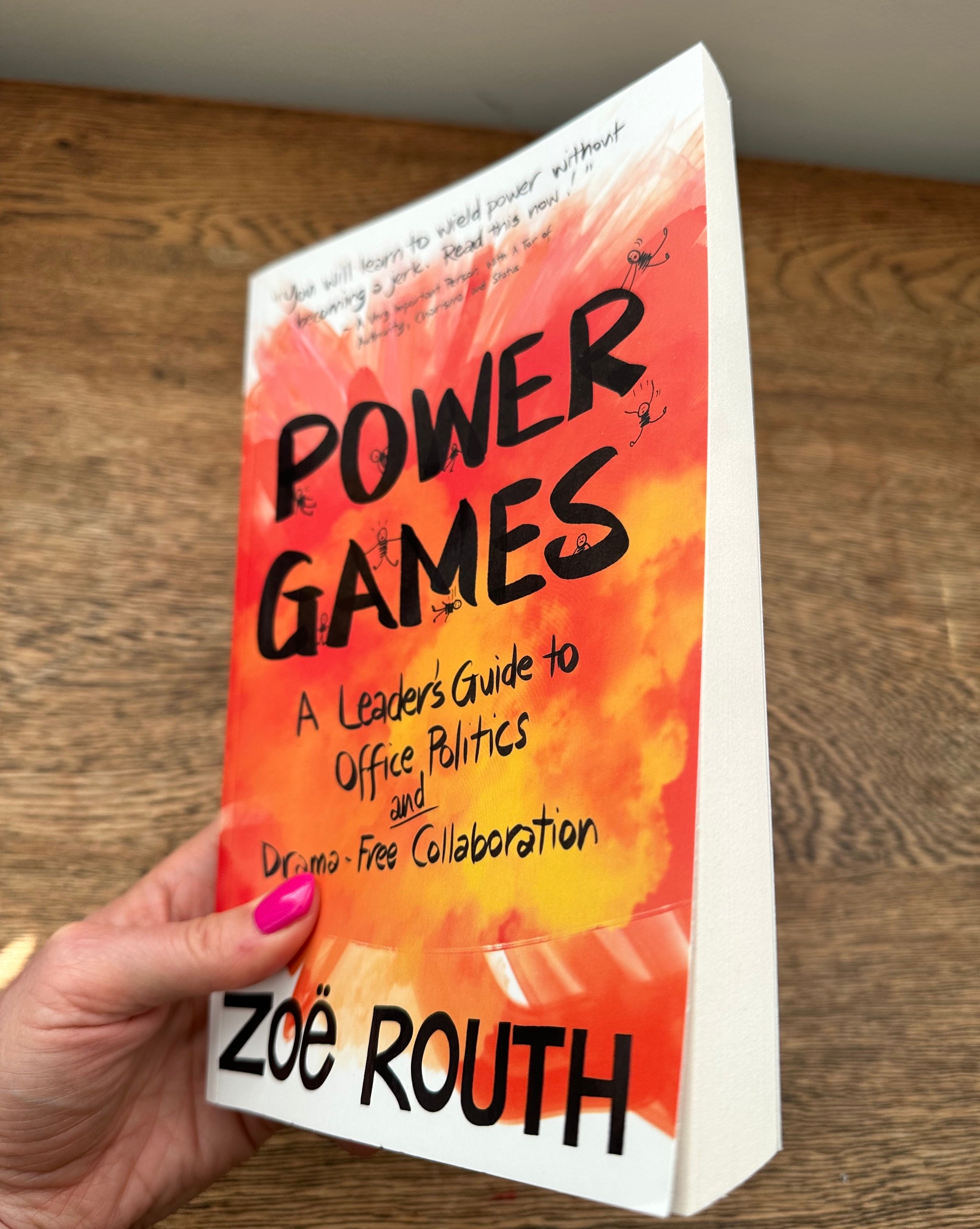 A hand with pink nail polish holds a paperback book titled 'Power Games' by Zoë Routh. The book cover features a vibrant orange and yellow abstract background with bold black text.