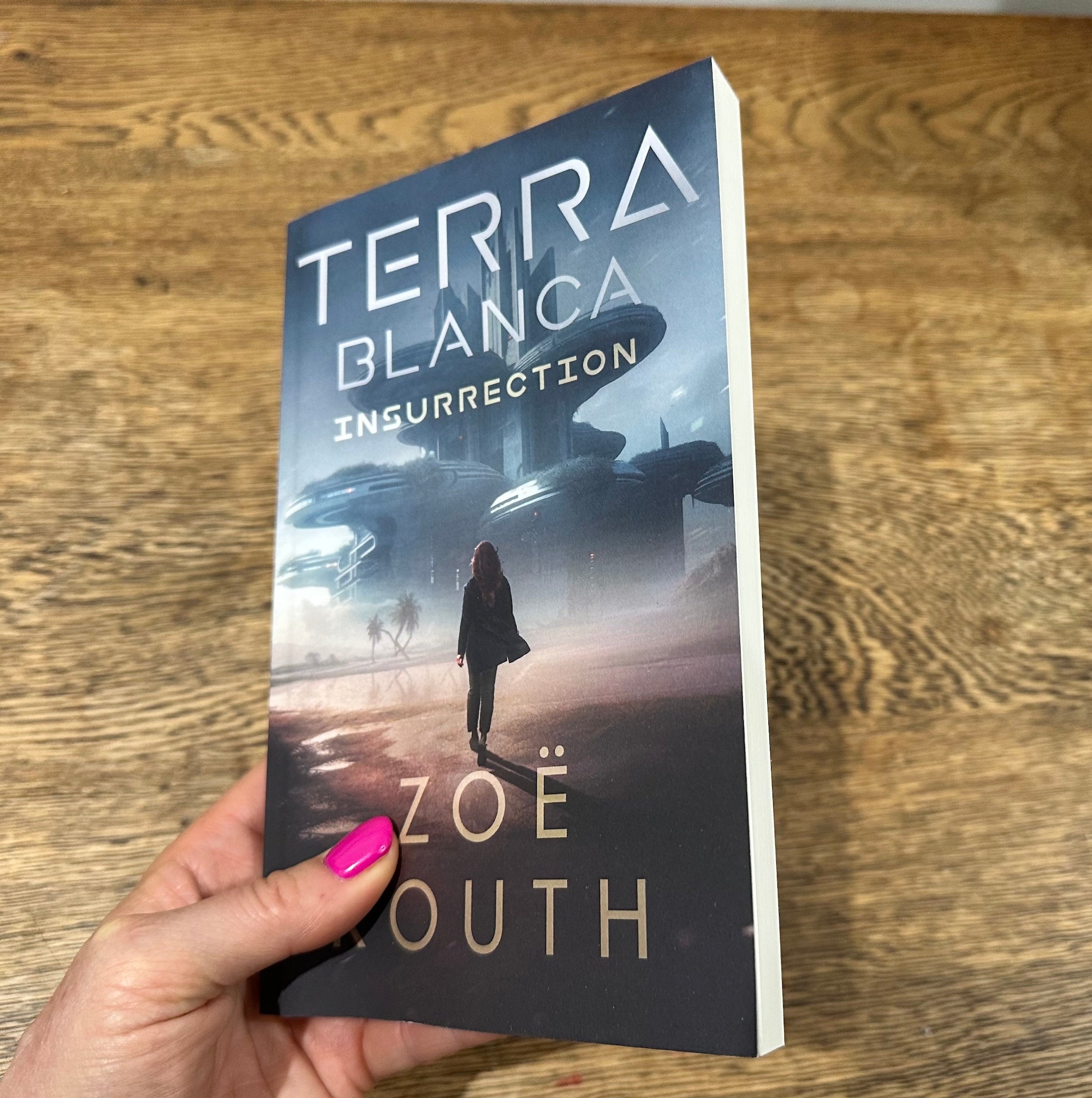A hand with bright pink nail polish holds a paperback book titled 'Terra Blanca: Insurrection' by Zoë Routh, showing the sci-fi cover art against a wooden tabletop background.