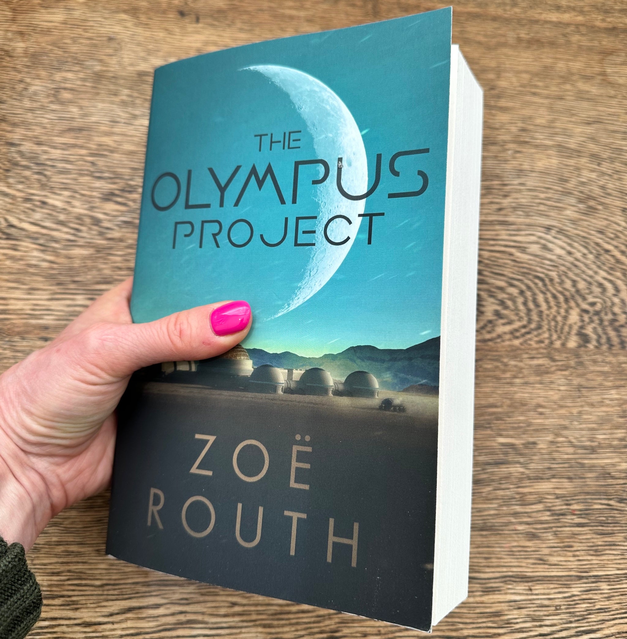 A hand with pink nail polish holds a paperback book titled 'The Olympus Project' by Zoë Routh, showing the front cover with a crescent moon and futuristic domes, set against a wooden tabletop background.